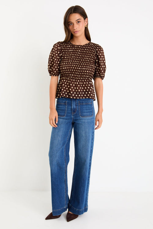 Model wearing a chocolate brown polka dot short sleeve top with a shirred bodice and peplum hem