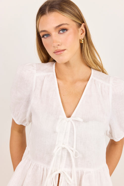 A woman wearing a white short-sleeve blouse with a deep V-neckline, gathered shoulders, and front tie details, photographed against a plain light background.