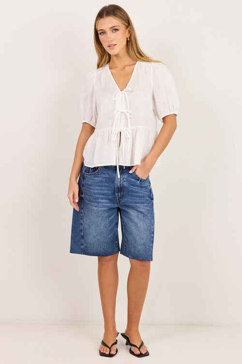 A woman modeling a white short-sleeve tie-front blouse and blue denim shorts against a plain off-white studio background.