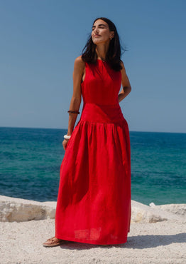 Woman in a red dress standing on a rocky beach with clear blue sky and ocean.