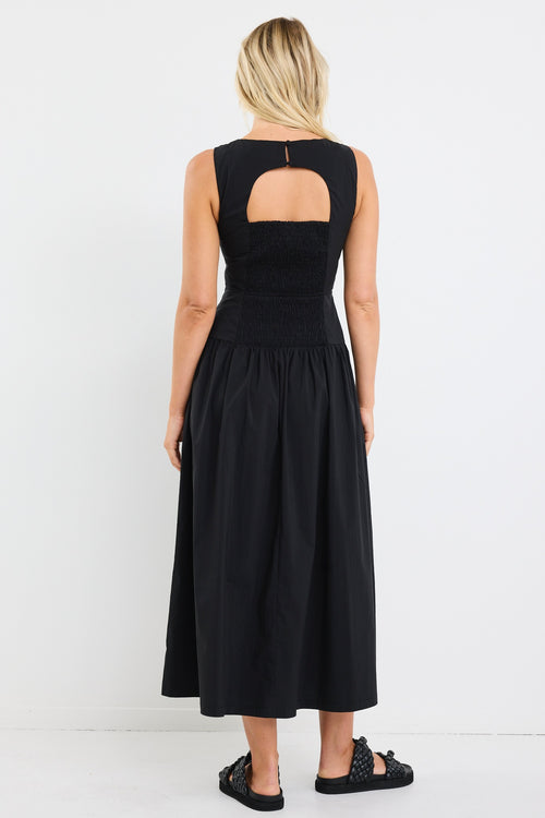 Woman wearing a sleeveless black midi dress with a fitted bodice and gathered skirt, styled with black braided sandals and gold hoop earrings.







