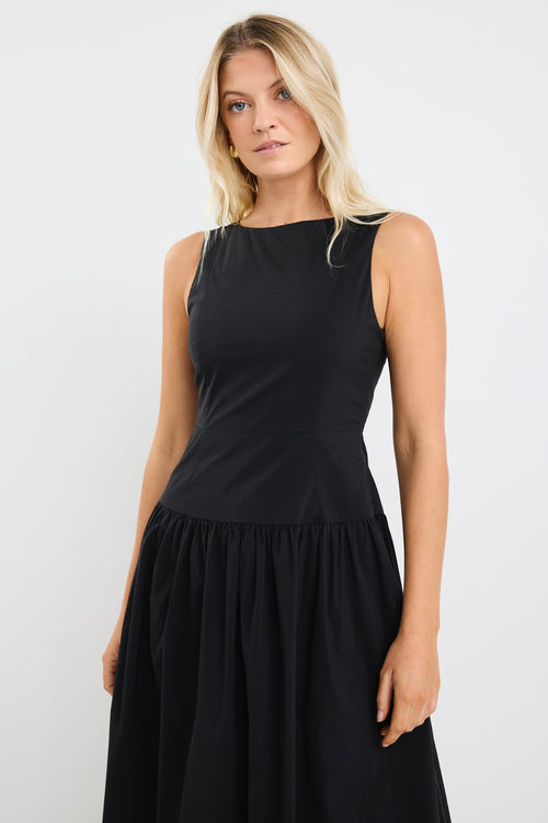 Woman wearing a sleeveless black midi dress with a fitted bodice and gathered skirt, styled with black braided sandals and gold hoop earrings.







