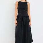 Woman wearing a sleeveless black midi dress with a fitted bodice and gathered skirt, styled with black braided sandals and gold hoop earrings.







