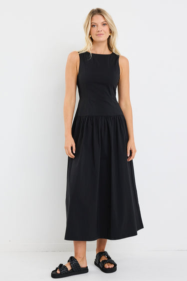 Woman wearing a sleeveless black midi dress with a fitted bodice and gathered skirt, styled with black braided sandals and gold hoop earrings.







