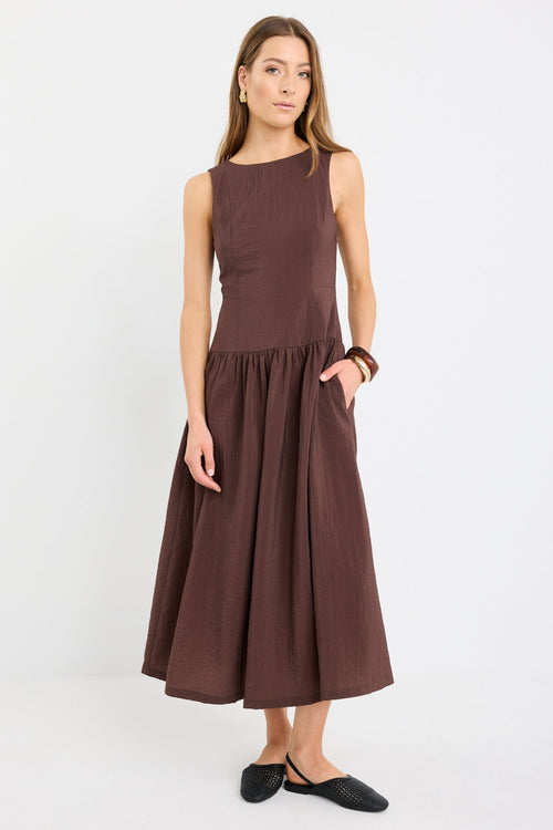 Woman wearing a chocolate brown sleeveless maxi dress with a structured bodice and flowing skirt, styled with black flat shoes