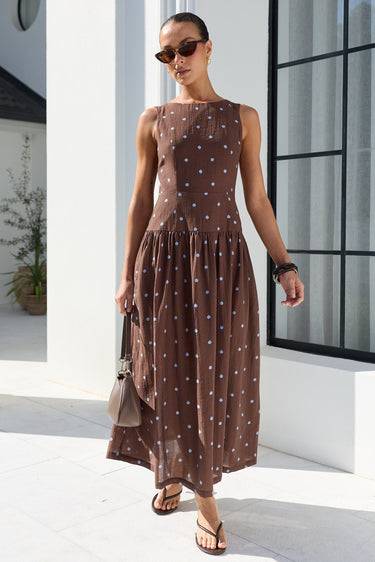 A woman wearing a sleeveless brown midi dress with a white polka dot pattern, styled with sunglasses, a small handbag, and sandals, walking outdoors in front of a white building with a large black-framed window.