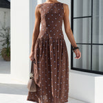 A woman models a sleeveless brown maxi dress with a blue polka dot pattern, walking outdoors on a white tiled patio next to a white building with a large black-framed window.