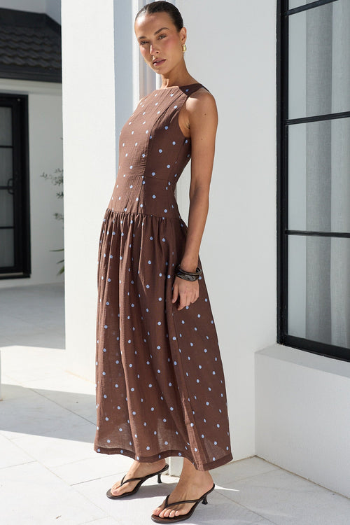 A woman modeling a sleeveless brown maxi dress with a white polka dot pattern, standing in a bright, modern white architectural setting with a large black-framed window.