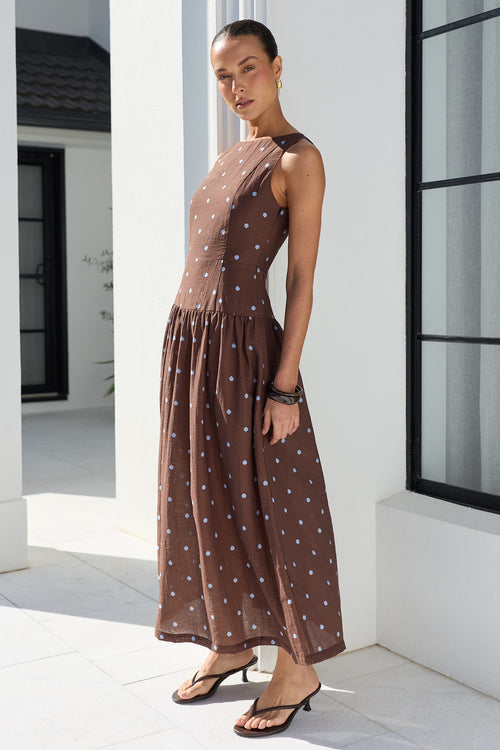A woman modeling a sleeveless brown maxi dress with a blue polka dot pattern, standing in a bright, modern white architectural setting with a large black-framed window.
