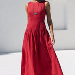 A woman wearing a sleeveless red midi dress and sunglasses walks along a white tiled poolside path next to a white stucco building.