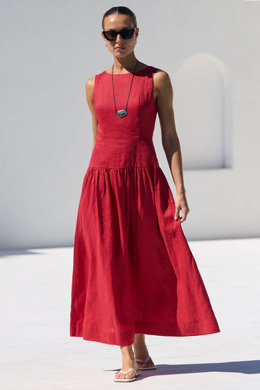 A woman wearing a sleeveless red midi dress and sunglasses walks along a white tiled poolside path next to a white stucco building.