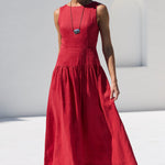 A woman wearing a sleeveless red midi dress, sunglasses, and a long necklace walks by a white poolside next to a white building with arched windows.