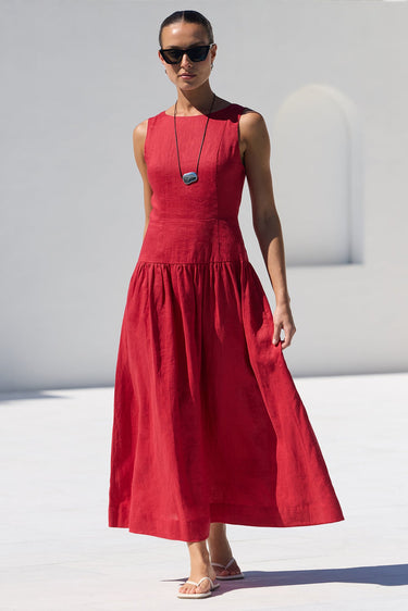A woman wearing a sleeveless red midi dress, sunglasses, and a long necklace walks by a white poolside next to a white building with arched windows.