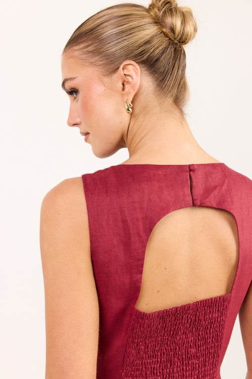 A woman shown from the back wearing a sleeveless burgundy dress with a large open back and smocked fabric panel, photographed against a plain light background.