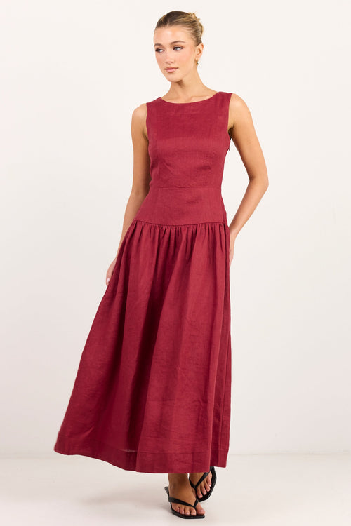 A woman modeling a sleeveless, floor-length maroon dress with a structured bodice and a flowing skirt, paired with black strappy sandals, against a plain white studio background.