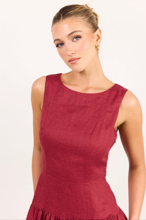 A woman wearing a sleeveless, scoop-neck dress in a textured deep burgundy fabric, photographed against a clean off-white studio background.