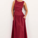 A woman modeling a sleeveless, floor-length maroon dress with side pockets, standing against a plain white studio background.