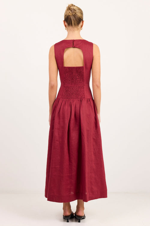 A woman viewed from the back wearing a sleeveless burgundy maxi dress with a smocked bodice and a keyhole cutout at the back, paired with black heels, against a plain white studio background.