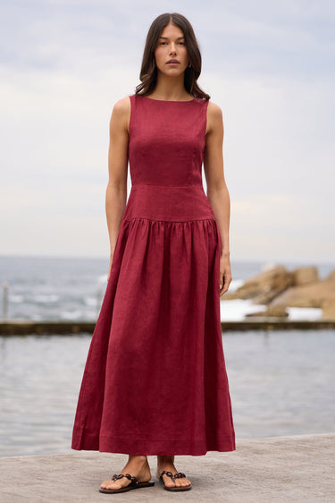 A woman stands outdoors by the water wearing a sleeveless, high-waisted maxi dress in a deep burgundy red. The dress has a structured bodice and a flowing skirt, paired with brown sandals.
