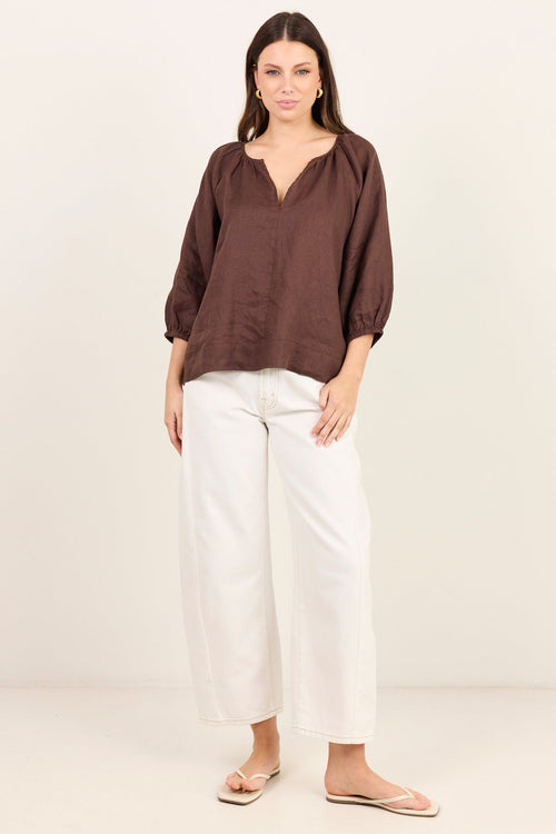 A woman modeling a brown long-sleeve blouse with a split neckline and white wide-leg cropped pants, standing against a plain off-white studio background.