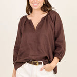 A woman modeling a brown long-sleeve blouse with a split neckline and gathered sleeves, paired with white pants and a gold belt, against a plain off-white background.