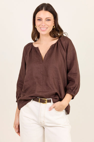 A woman modeling a brown long-sleeve blouse with a split neckline and gathered sleeves, paired with white pants and a gold belt, against a plain off-white background.