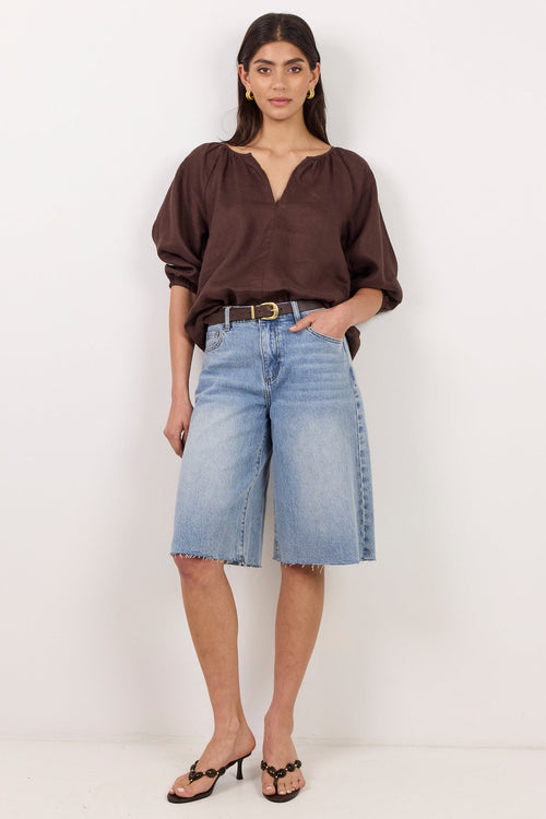 A woman modeling a dark brown long-sleeve blouse and light-wash denim cropped shorts with a black belt, standing against a plain white studio background.