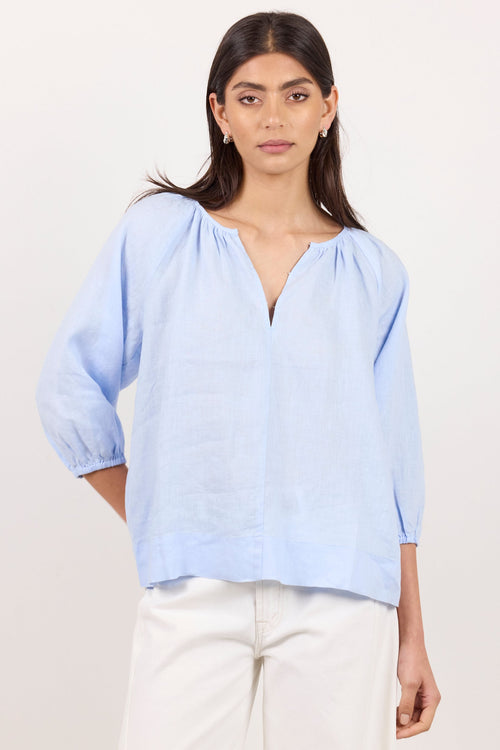 A woman modeling a light blue linen blouse with a split V-neck and gathered sleeves, paired with white wide-leg pants, against a plain off-white studio background.
