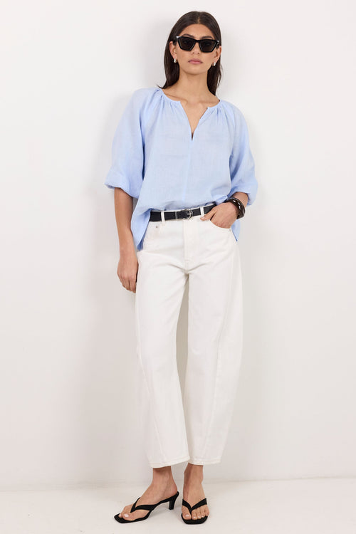 A woman modeling a light blue short-sleeve blouse and white cropped trousers with a black belt, styled with black sandals and sunglasses against a plain white studio background.