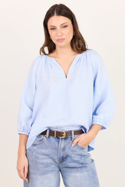 A woman modeling a light blue, loose-fitting blouse with voluminous three-quarter sleeves and a gathered V-neckline, paired with light-wash blue jeans and a brown belt, against a plain off-white studio background.