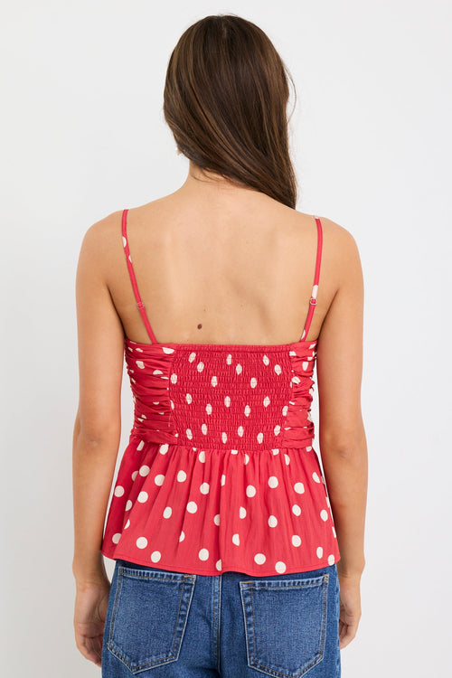  Woman wearing a red polka dot ruched cami top with thin straps and a peplum hem, styled with blue jeans against a plain white background.