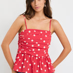 Woman wearing a red polka dot ruched cami top with thin straps and a peplum hem, styled with blue jeans against a plain white background.