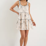 Woman wearing a sleeveless, tiered mini dress with strappy sleeves and a floral sketch print on an ivory background.