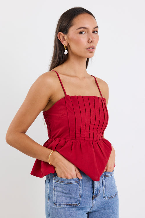 Woman wearing a plum-red pleated bodice peplum top with thin straps, paired with blue jeans and gold jewellery, standing against a white background.