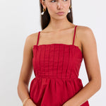 Woman wearing a plum-red pleated bodice peplum top with thin straps, paired with blue jeans and gold jewellery, standing against a white background.