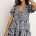 Woman wearing a navy gingham short sleeve dress with puff sleeves, a tie-front bodice, and tiered skirt, paired with white sneakers.