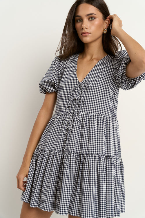 Woman wearing a navy gingham short sleeve dress with puff sleeves, a tie-front bodice, and tiered skirt, paired with white sneakers.
