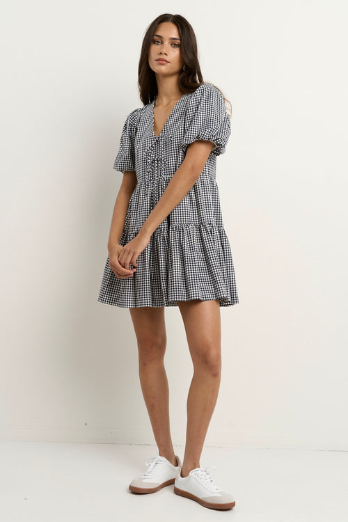 Woman wearing a navy gingham short sleeve dress with puff sleeves, a tie-front bodice, and tiered skirt, paired with white sneakers.