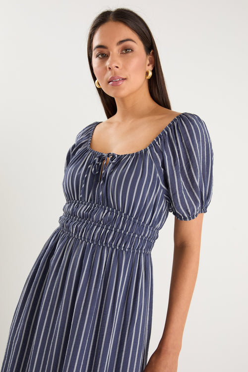 Woman wearing a navy maxi dress with vertical white stripes, short puff sleeves, a smocked waist, and side slit detail.
