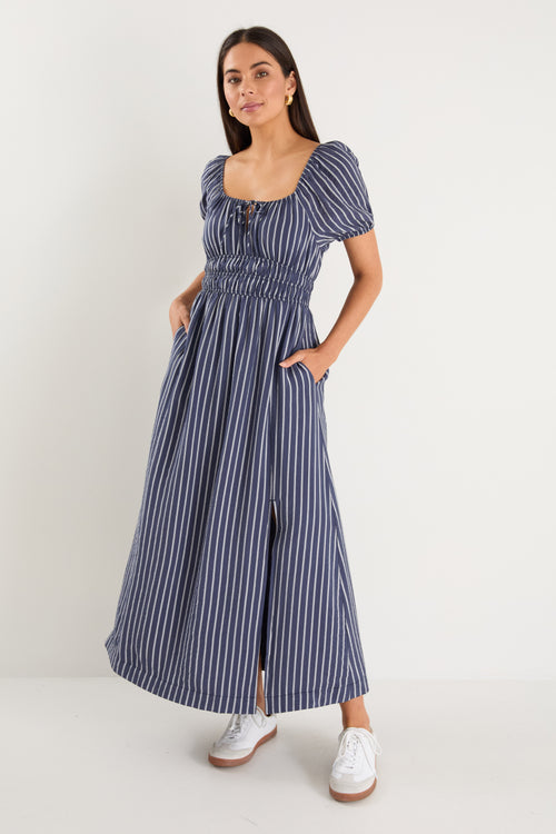 Woman wearing a navy maxi dress with vertical white stripes, short puff sleeves, a smocked waist, and side slit detail.