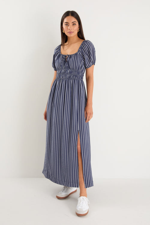 Woman wearing a navy maxi dress with vertical white stripes, short puff sleeves, a smocked waist, and side slit detail.