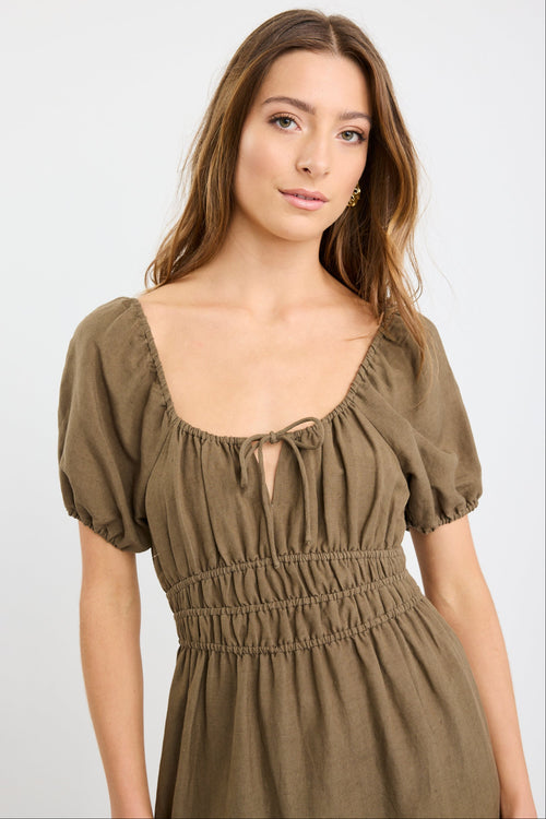 Woman wearing a khaki short sleeve linen blend midi dress with a shirred waist, tie neckline, and side split, styled with white sneakers.