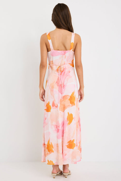 Woman wearing a strappy maxi dress with an abstract pink and orange sunset print, cut on the bias for a floaty and feminine silhouette.