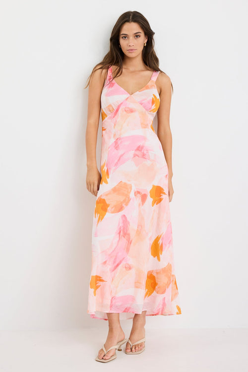 Woman wearing a strappy maxi dress with an abstract pink and orange sunset print, cut on the bias for a floaty and feminine silhouette.