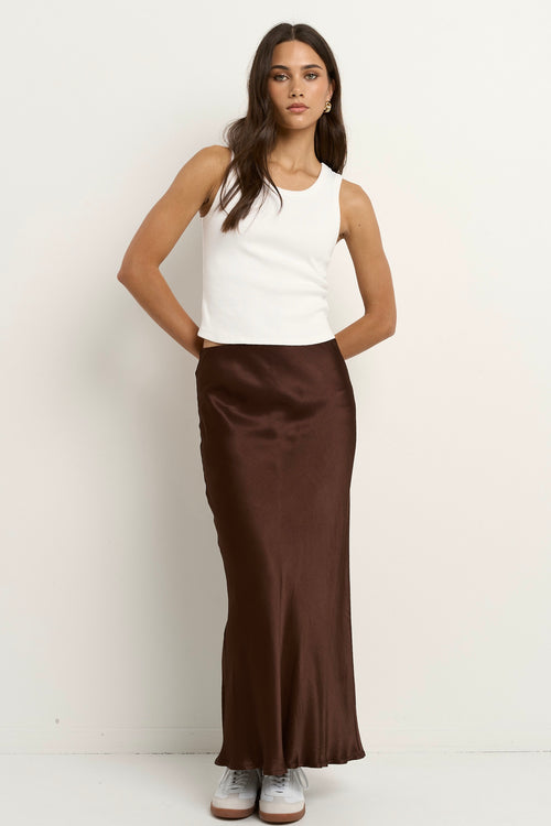 Woman wearing a chocolate brown satin midi skirt with a white tank and sneakers.