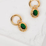 A pair of gold huggie hoop earrings with oval green gemstone charms, displayed on a textured white surface.