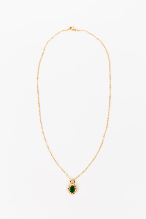 A delicate gold chain necklace with a small oval pendant featuring a deep green stone surrounded by a gold border, displayed against a plain white background.