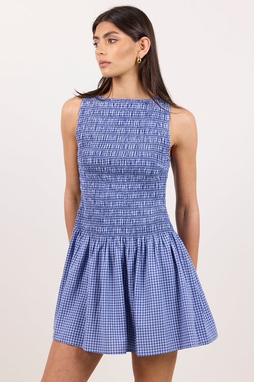A woman modeling a sleeveless smocked mini dress with a blue and white gingham pattern, photographed against a plain light beige studio background.