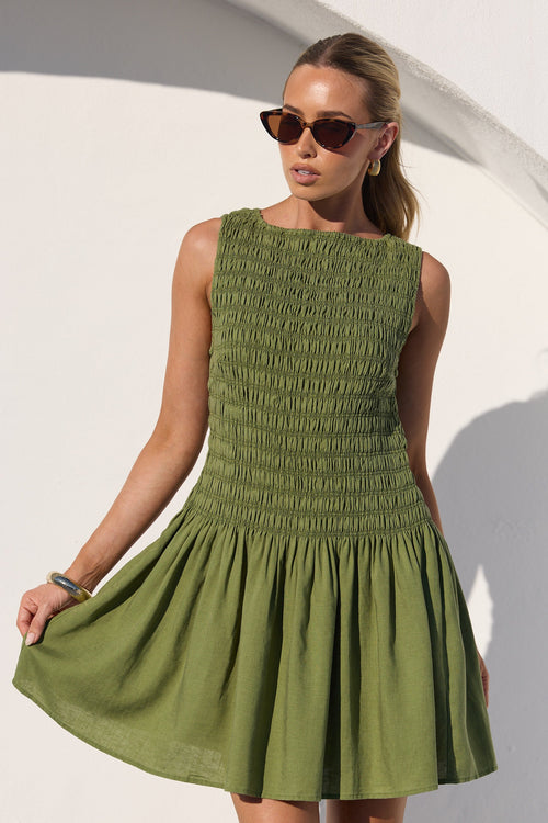 A woman modeling a sleeveless olive green dress with a smocked bodice and flared skirt, standing against a white wall with a strong shadow.