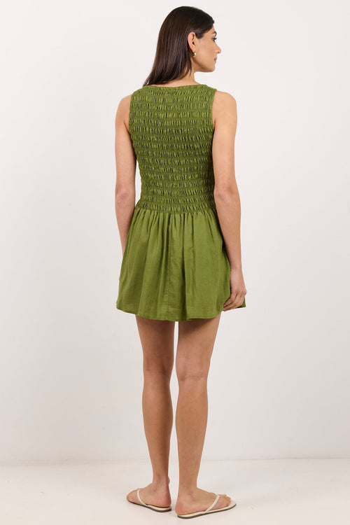 A woman stands with her back to the camera wearing a sleeveless olive green mini dress with a smocked bodice and a flared skirt, paired with beige flat sandals, against a plain white studio background.